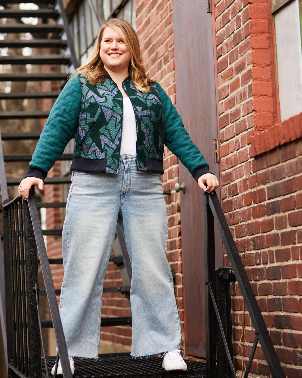 Woman wearing a green patterned jacket and blue jeans standing on a staircase with a brick wall background. This jacket is the Cashmerette Club Kimball Bomber Jacket sewing pattern.