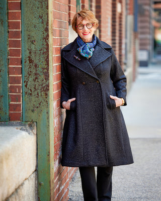 Woman wearing a dark coat and colorful scarf leaning against a brick wall. This coat is the Savin Coat sewing pattern from Cashmerette.