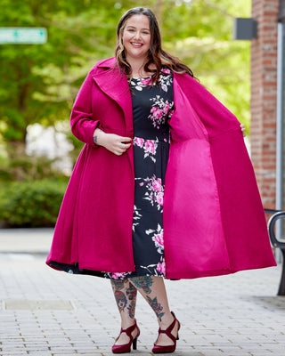 Woman wearing a bright pink coat over a floral dress on a street. This coat is the Savin Coat sewing pattern from Cashmerette.
