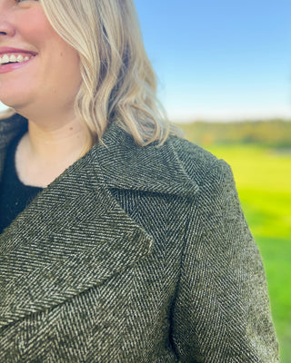 Person wearing a green herringbone coat with a blurred natural background. This coat is the Savin Coat sewing pattern from Cashmerette.