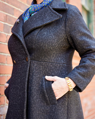 Person wearing a dark gray coat with a brick wall background. This coat is the Savin Coat sewing pattern from Cashmerette.