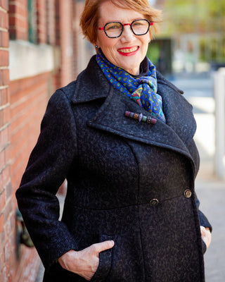 Woman wearing a dark coat and colorful scarf standing against a brick wall. This coat is the Savin Coat sewing pattern from Cashmerette.