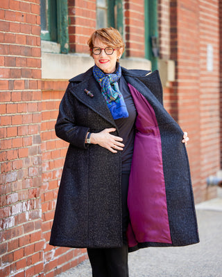 Person wearing a long dark coat with a colorful scarf against a brick wall. This coat is the Savin Coat sewing pattern from Cashmerette.