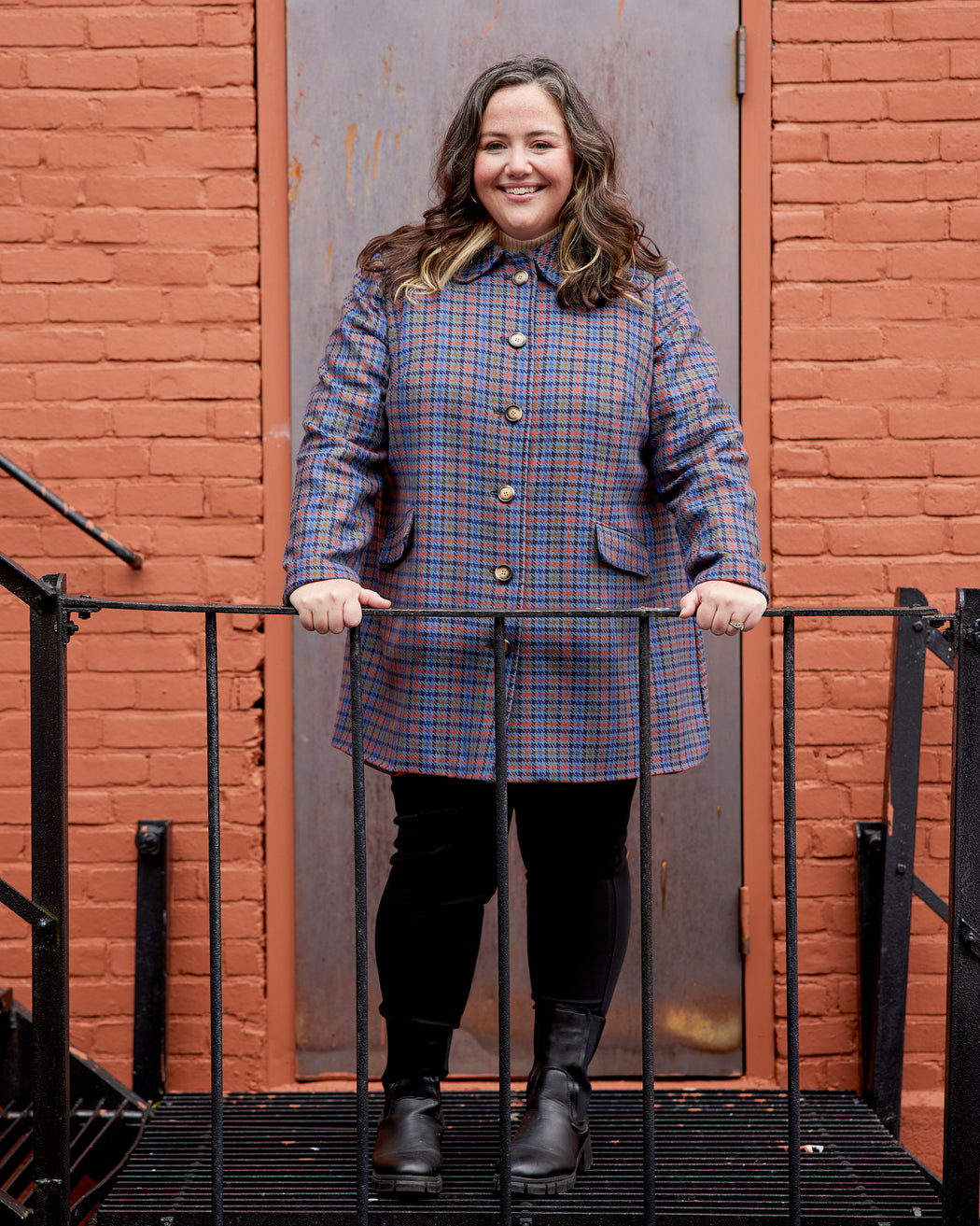 Woman wearing a plaid coat standing against a red brick wall. This coat is the Newcomb Car Coat sewing pattern from Cashmerette.