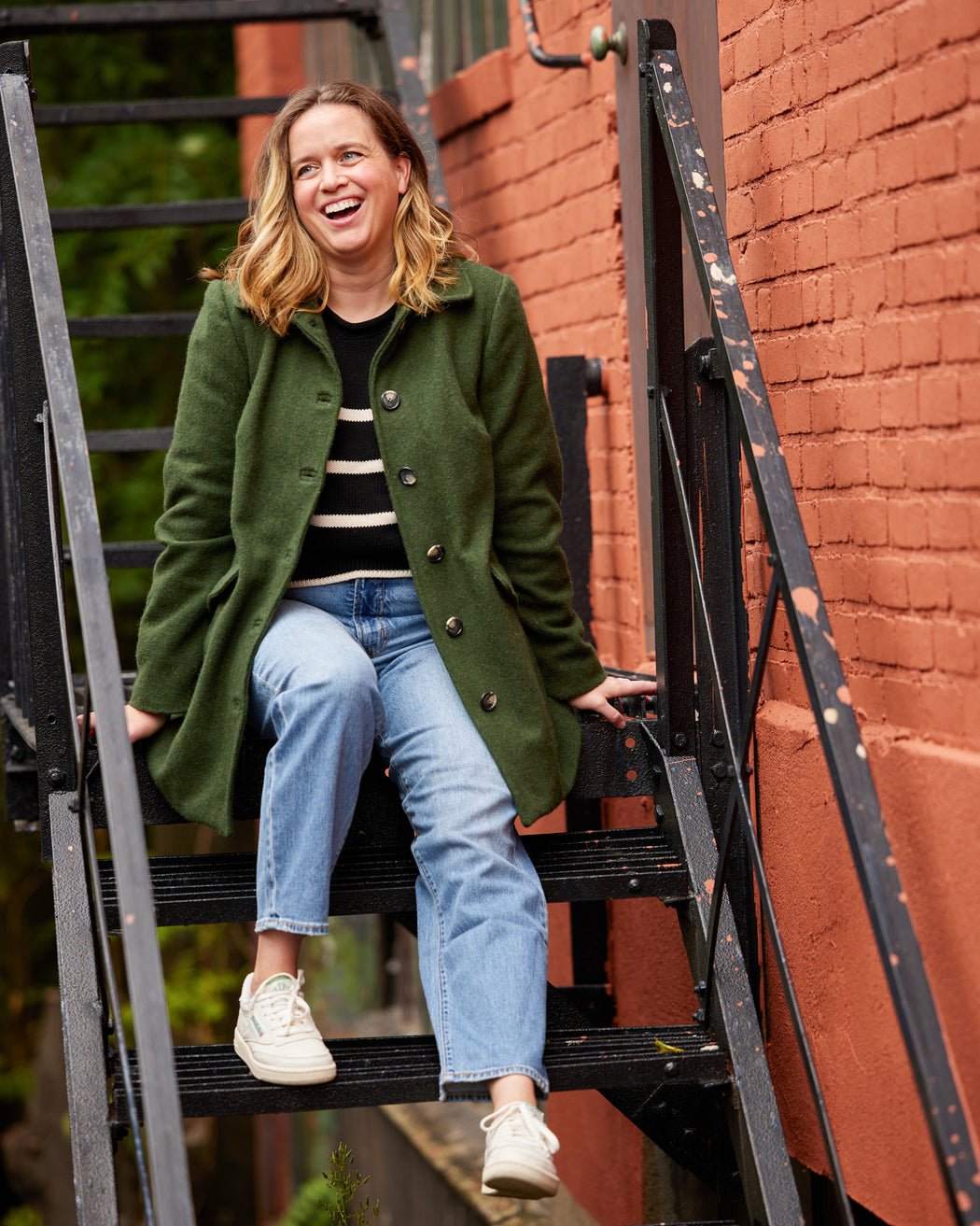 Woman in a green coat sitting on a staircase with a brick wall background. This coat is the Newcomb Car Coat sewing pattern from Cashmerette.