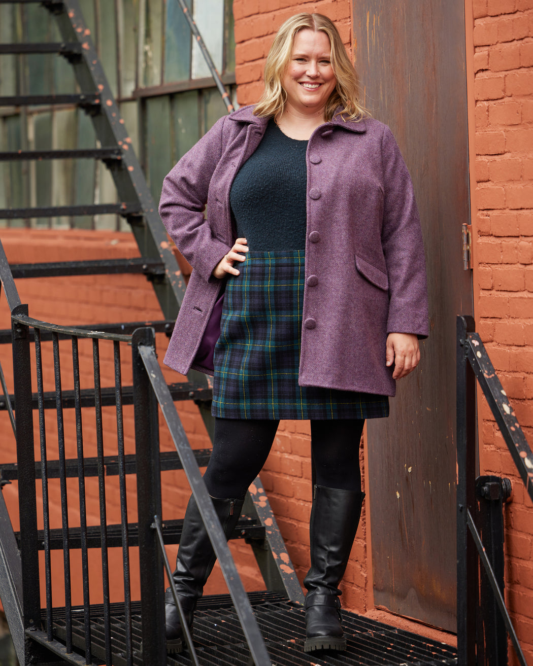 Woman wearing a purple coat, black dress, and plaid skirt standing in front of a brick wall. This coat is the Newcomb Car Coat sewing pattern from Cashmerette.