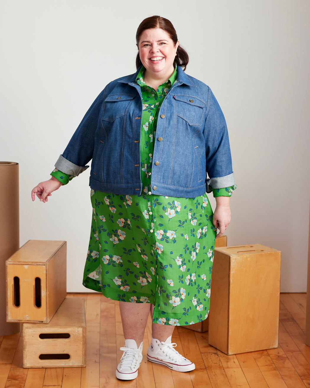 Woman wearing a blue denim jacket over a green floral dress standing in front of wooden boxes. This jacket is the Glover Jean Jacket sewing pattern from Cashmerette.