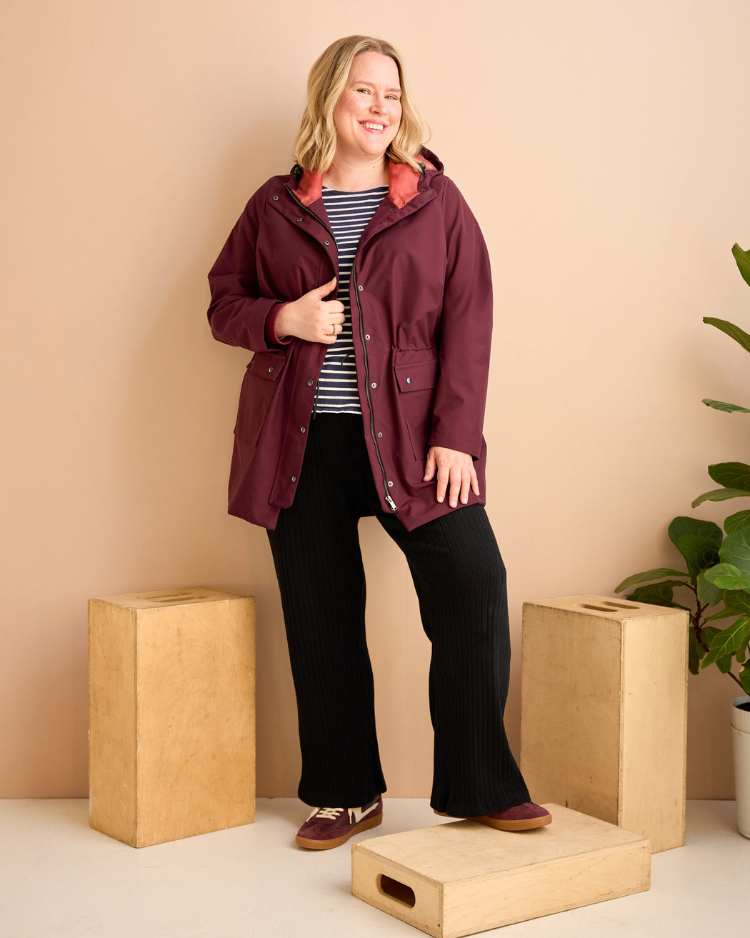 Woman wearing a burgundy jacket standing in a minimalistic setting with beige walls and wooden blocks. This is the Marston Raincoat sewing pattern from Cashmerette Club.