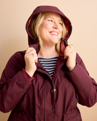 Woman wearing a maroon raincoat against a beige background. This is the Marston Raincoat sewing pattern from Cashmerette Club.