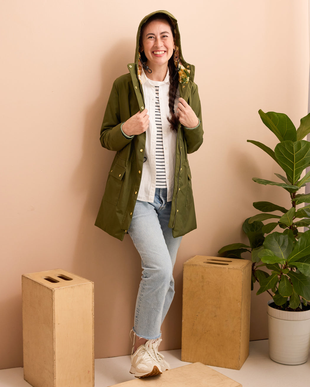 Woman wearing a green raincoat indoors with a beige wall and plant in the background. This is the Marston Raincoat sewing pattern from Cashmerette Club.