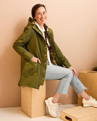 Woman wearing a green rain jacket sitting on a cardboard box against a beige wall.  This is the Marston Raincoat sewing pattern from Cashmerette Club.