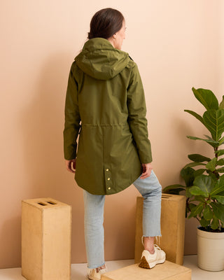 Person wearing a green raincoat standing against a beige wall with a plant in the background. This is the Marston Raincoat sewing pattern from Cashmerette Club.