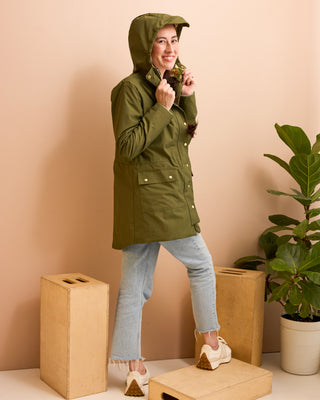 Person wearing a green raincoat standing in a minimalistic room with a plant and boxes. This is the Marston Raincoat sewing pattern from Cashmerette Club.