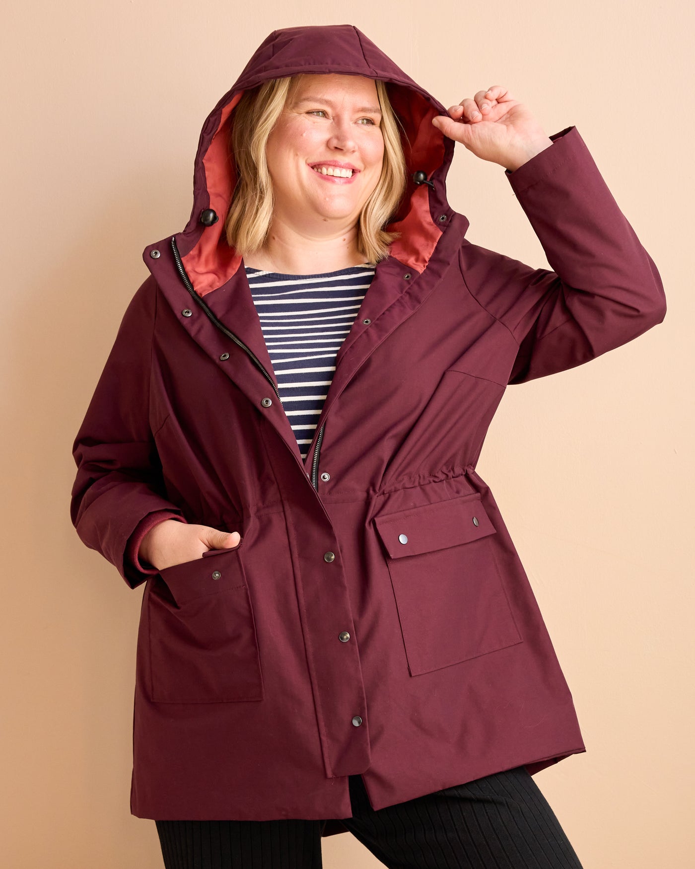Woman wearing a burgundy raincoat with a hood on a beige background. This is the Marston Raincoat sewing pattern from Cashmerette Club.