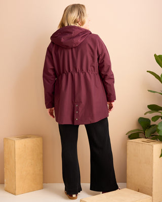 Person wearing a burgundy raincoat standing against a beige wall with a plant in the background. This is the Marston Raincoat sewing pattern from Cashmerette Club.