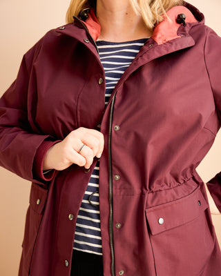 Person wearing a burgundy raincoat with a striped shirt underneath on a beige background. This is the Marston Raincoat sewing pattern from Cashmerette Club.