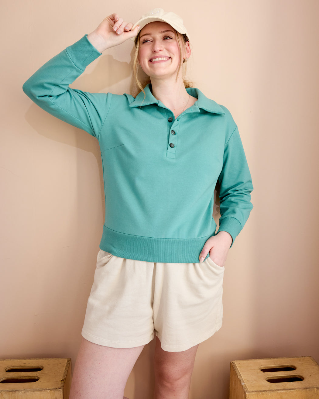Woman wearing a teal long-sleeve polo shirt and beige shorts against a beige background. This is the Brookhaven Sweatshirt sewing pattern from Cashmerette.