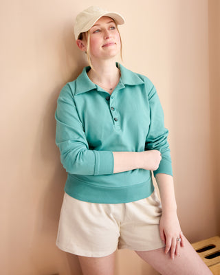 Woman wearing a teal sweater and beige shorts against a beige wall. This is the Brookhaven Sweatshirt sewing pattern from Cashmerette.