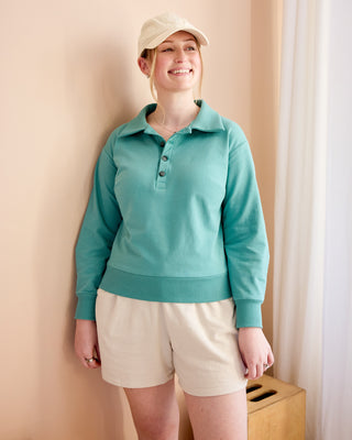Person wearing a teal long-sleeve shirt and beige shorts against a beige wall. This is the Brookhaven Sweatshirt sewing pattern from Cashmerette.