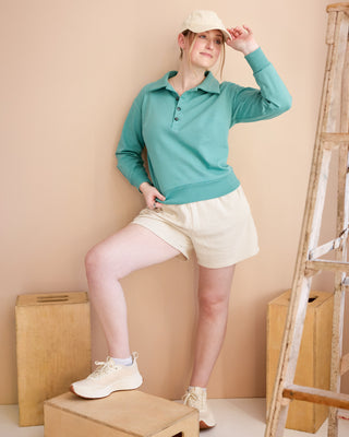 Person wearing a teal long-sleeve shirt and white shorts, standing on a wooden box against a beige wall. This is the Brookhaven Sweatshirt sewing pattern from Cashmerette.