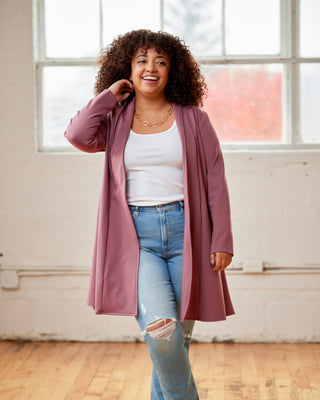 Woman wearing a long pink coat over a white top and blue jeans in a bright room. This cardigan is the Westmore Cardigan sewing pattern from Cashmerette.