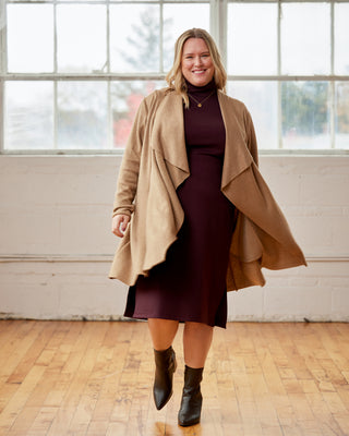 Woman wearing a beige coat over a dark dress with black boots in a room with large windows. This cardigan is the Westmore Cardigan sewing pattern from Cashmerette.