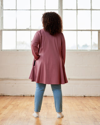Person wearing a pink dress and blue jeans standing in a room with large windows. This cardigan is the Westmore Cardigan sewing pattern from Cashmerette.