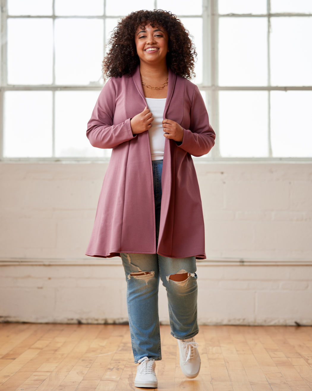 Woman wearing a pink cardigan over a white shirt and blue jeans, standing in a room with large windows. This cardigan is the Westmore Cardigan sewing pattern from Cashmerette.