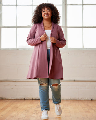Woman wearing a pink cardigan over a white shirt and blue jeans, standing in a room with large windows. This cardigan is the Westmore Cardigan sewing pattern from Cashmerette.