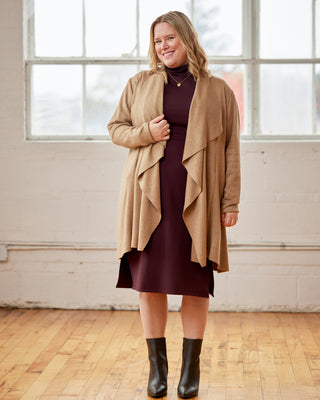 Woman wearing a beige cardigan over a dark dress with black boots in a room with large windows. This cardigan is the Westmore Cardigan sewing pattern from Cashmerette.