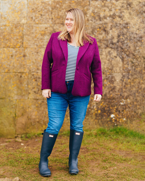 Woman wearing a purple jacket, striped shirt, blue jeans, and navy boots standing in front of a stone wall. This blazer is the Auburn Blazer sewing pattern from Cashmerette.