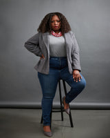 Woman sitting on a stool wearing a gray cardigan, white shirt, blue jeans, and gray shoes against a gray background. This blazer is the Auburn Blazer sewing pattern from Cashmerette.