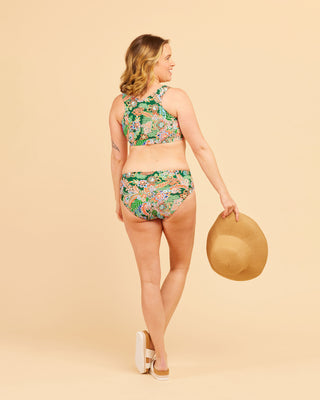 Woman wearing a floral bikini holding a hat on a beige background. This swimsuit is the Marblehead Swimsuit sewing pattern from Cashmerette.