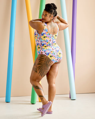 Woman wearing a colorful floral swimsuit standing against a beige background with rainbow-colored cylindrical objects. This is the Truro Swimsuit sewing pattern from Cashmerette.