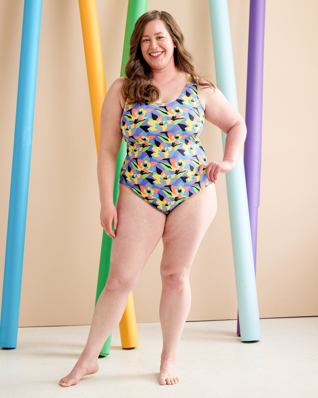 Woman wearing a colorful patterned swimsuit standing against a beige background with multicolored cylindrical props. This is the Truro Swimsuit sewing pattern from Cashmerette.