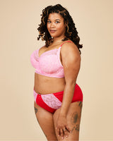 Woman wearing a pink lace bralette and red high-waisted underwear on a beige background. This bra is the Willowdale Bra sewing pattern from Cashmerette.