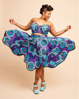 Woman twirling in a colorful purple and blue dress with a pattern on a beige background. This bra is the Symmes Strapless Bra sewing pattern from Cashmerette.