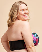 Woman wearing a strapless black top with colorful floral design underneath on a beige background. This bra is the Symmes Strapless Bra sewing pattern from Cashmerette.