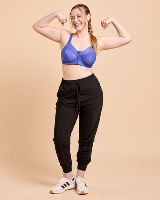 Woman flexing her muscles wearing a blue sports bra and black jogger pants on a beige background. This is the Cashmerette Somerville Sports Bra sewing pattern for big boobs.
