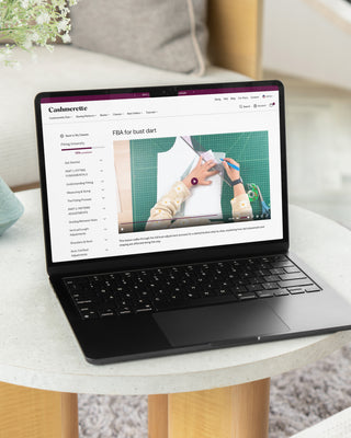 Laptop on a table with a website open on the screen, placed on a light-colored surface, showing a screenshot of a lesson from Cashmerette Fitting University.