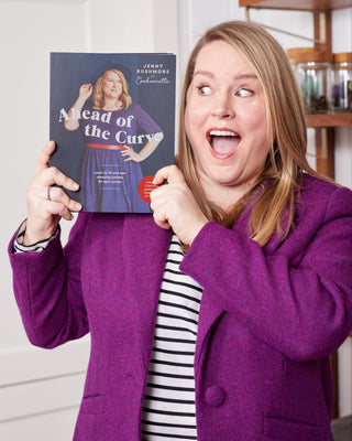 Person in a purple coat holding a book titled 'Ahead of the Curve' in a room with shelves.