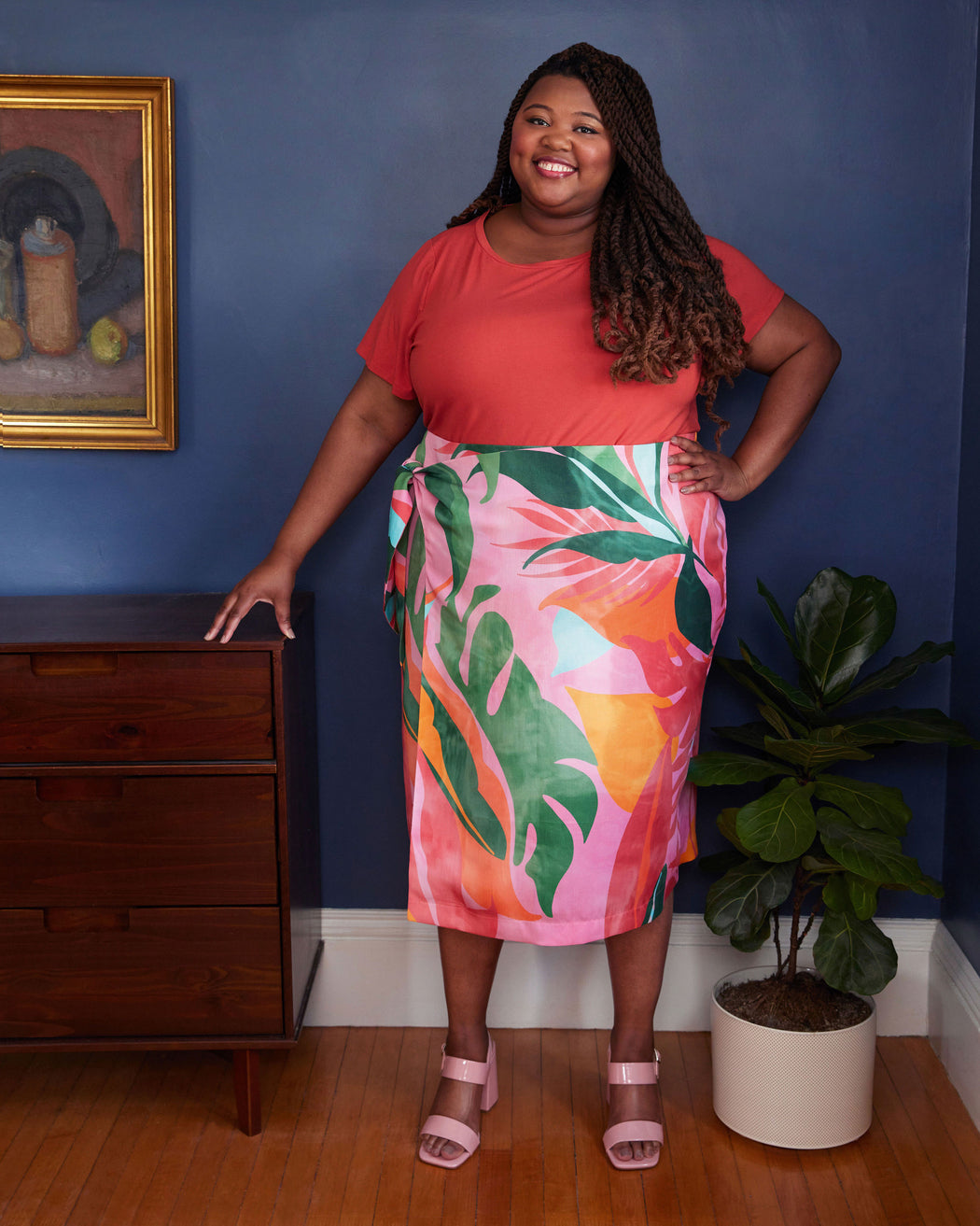 Woman in a red top and colorful skirt standing in a room with a blue wall, wooden dresser, and plant. This skirt is the Randolph Wrap Skirt sewing pattern from "Sewing the Curve" by Jenny Rushmore.