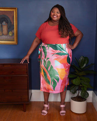 Woman in a red top and colorful skirt standing in a room with a blue wall, wooden dresser, and plant. This skirt is the Randolph Wrap Skirt sewing pattern from "Sewing the Curve" by Jenny Rushmore.