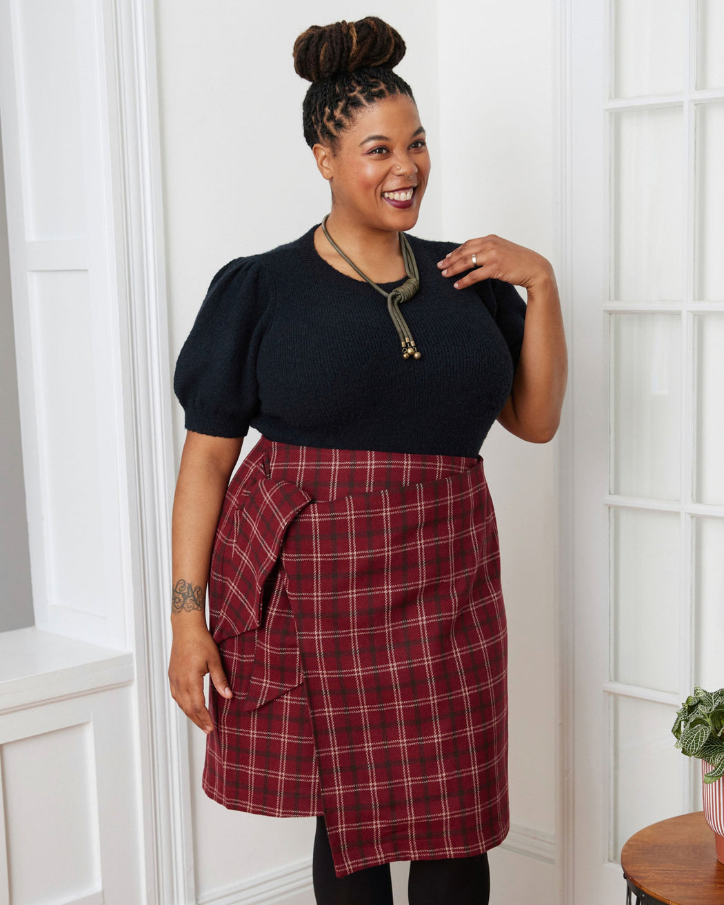Woman wearing a black top and red plaid skirt standing in a room with wooden flooring and a white wall. This skirt is the Randolph Wrap Skirt sewing pattern from "Sewing the Curve" by Jenny Rushmore.