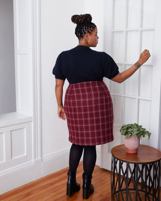 Person wearing a black top and red plaid skirt standing in a room with a plant on a table. This skirt is the Randolph Wrap Skirt sewing pattern from "Sewing the Curve" by Jenny Rushmore.