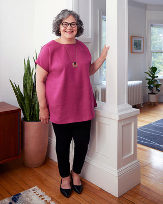Woman in a pink shirt standing in a home interior with plants and a radiator. This top is the Montvale Dress & Tunic sewing pattern from "Sewing the Curve" by Jenny Rushmore.