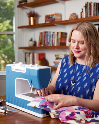 Woman using a blue sewing machine in a home setting with books and decor in the background.