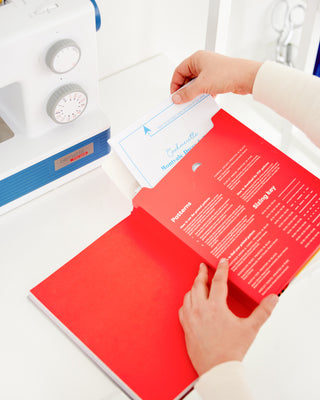 Person holding a red book with text and a white card on a sewing machine background. This book is Sewing the Curve by Jenny Rushmore.