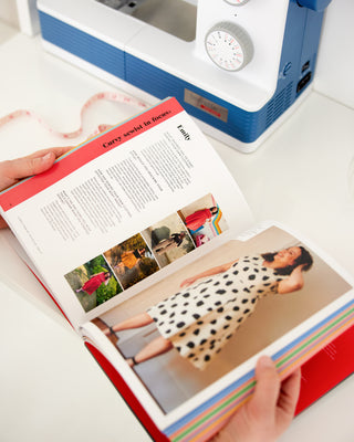 Open photo book with images and text, held by a person, with a sewing machine in the background. This book is Ahead of the Curve by Jenny Rushmore.