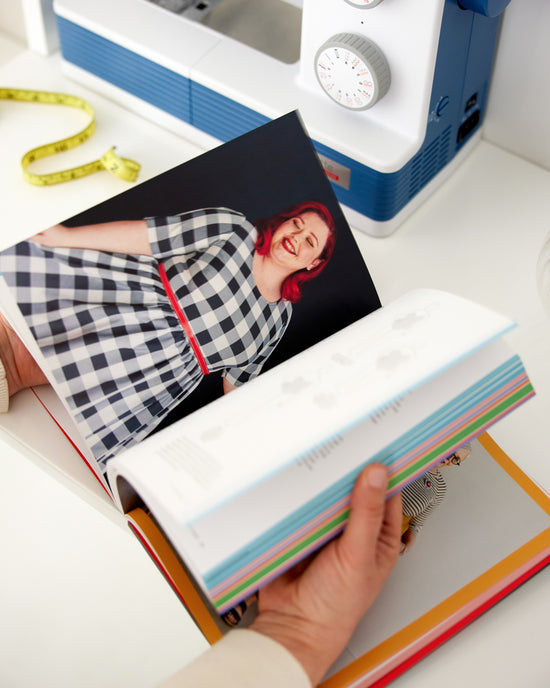 Person holding a book with a photo of a person on top, sewing machine in the background. This book is Ahead of the Curve by Jenny Rushmore.